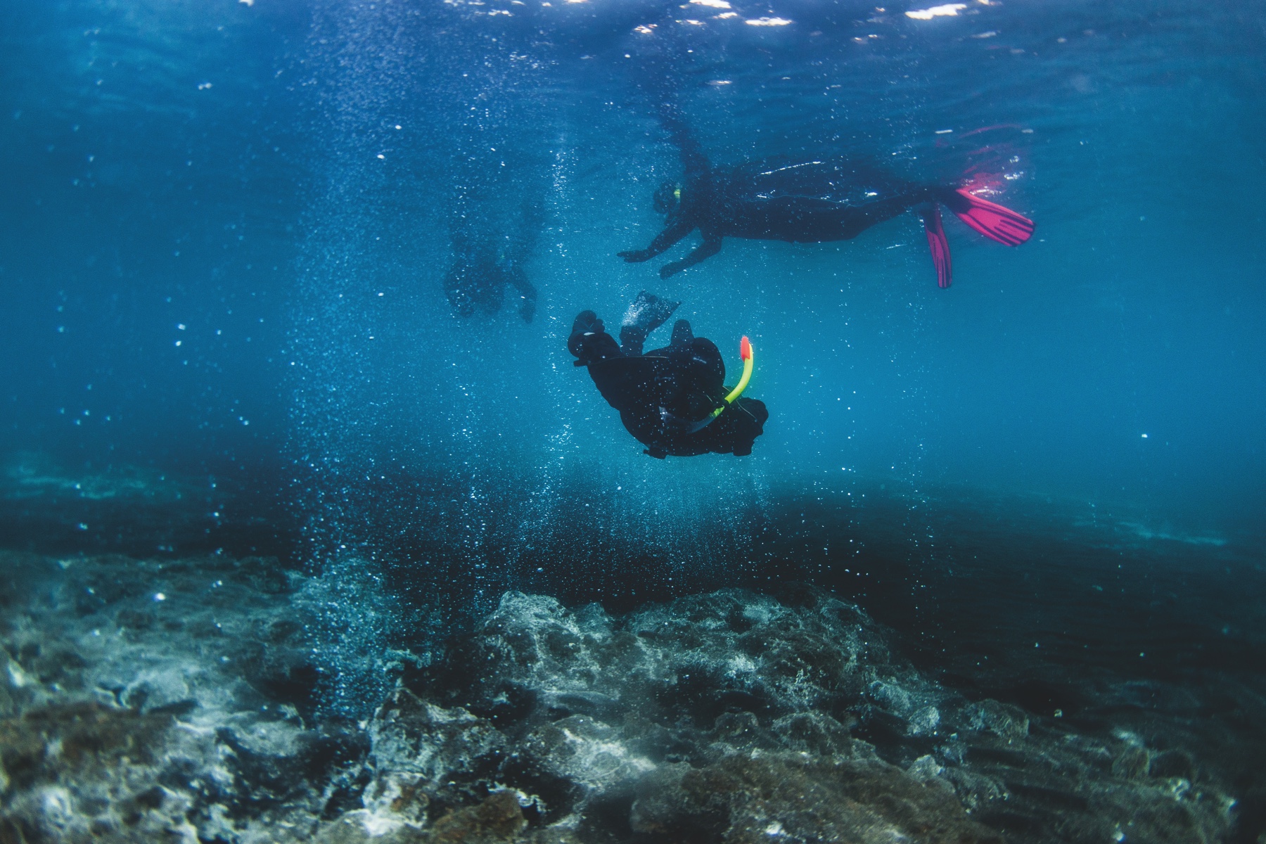 snorkeler diving towards camera between black sand bubbling hot water out of white rocks up to two snorkelers in Kleifarvatn