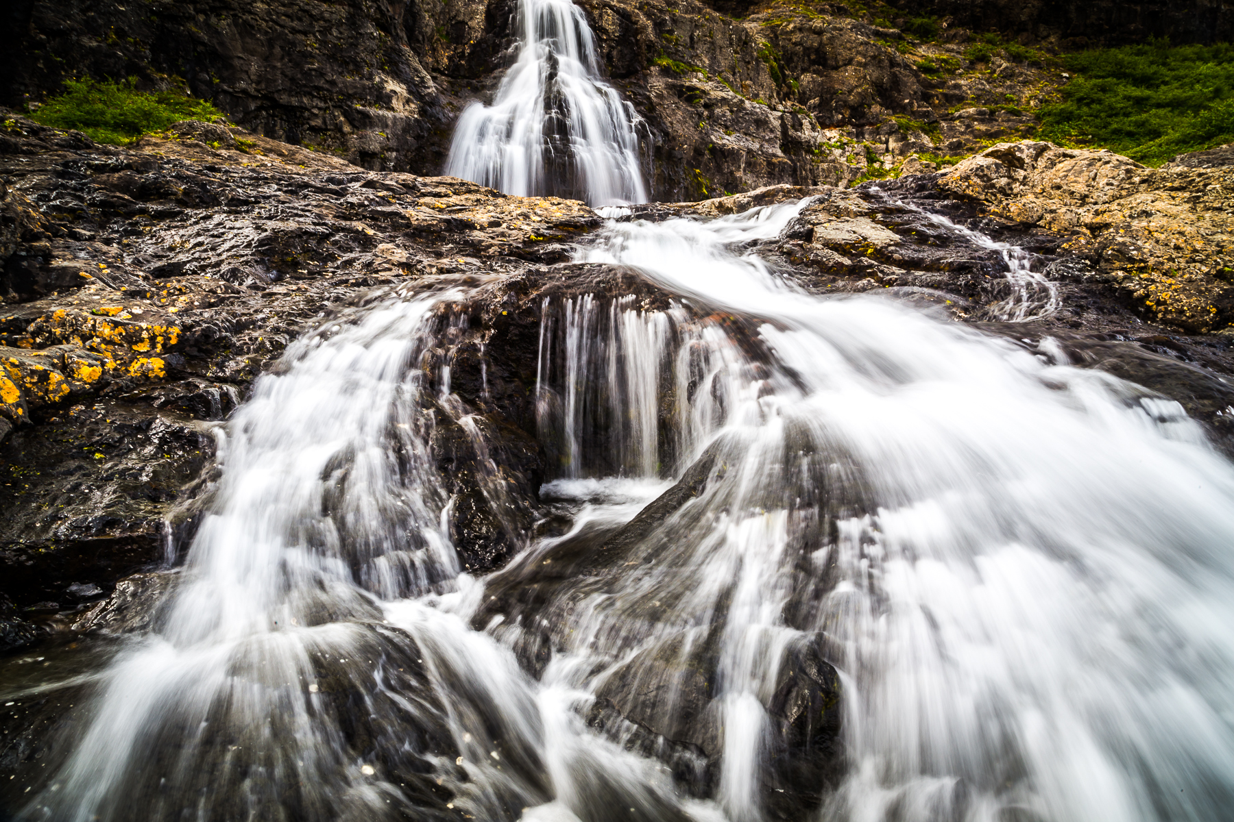 close-up of streaming spread out waterfall in two main steps bedded in dark icelandic rock covered in mustard yellow lychen