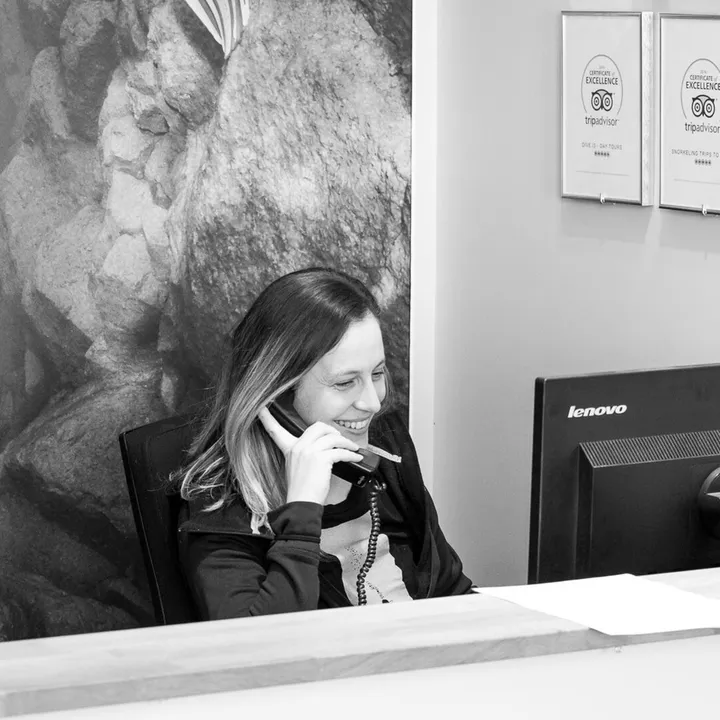 A smiling office angel answers the phone sitting at computer behind front desk in DIVE.IS shop in front of Silfra poster