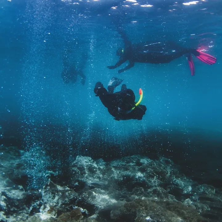 snorkeler diving towards camera between black sand bubbling hot water out of white rocks up to two snorkelers in Kleifarvatn