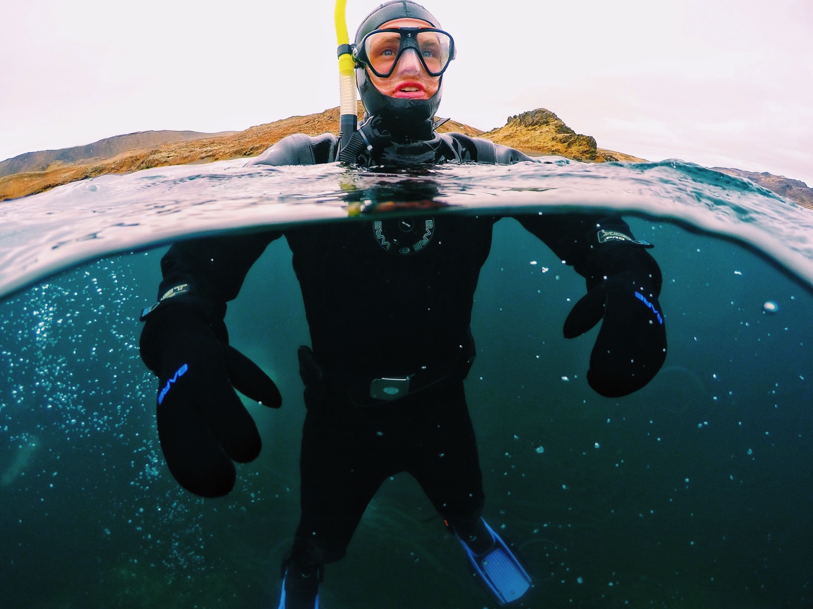 close-up split shot of snorkeler floating upright in lake Kleifarvatn surrounded by bubbles in the water before rocky shore