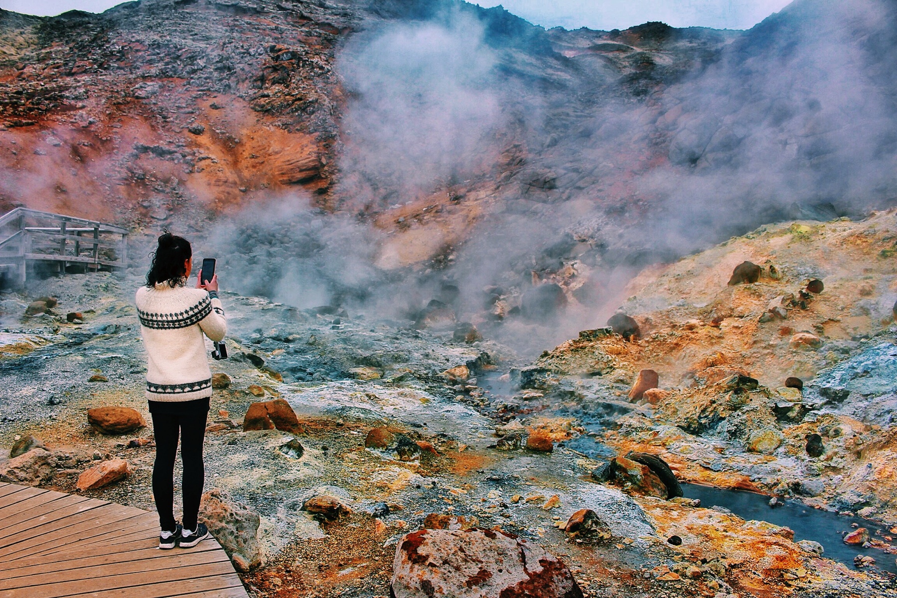 girl with white Icelandic sweater taking phone picture of steaming yellow sulfuric rocky stream at Krýsuvík geothermal site