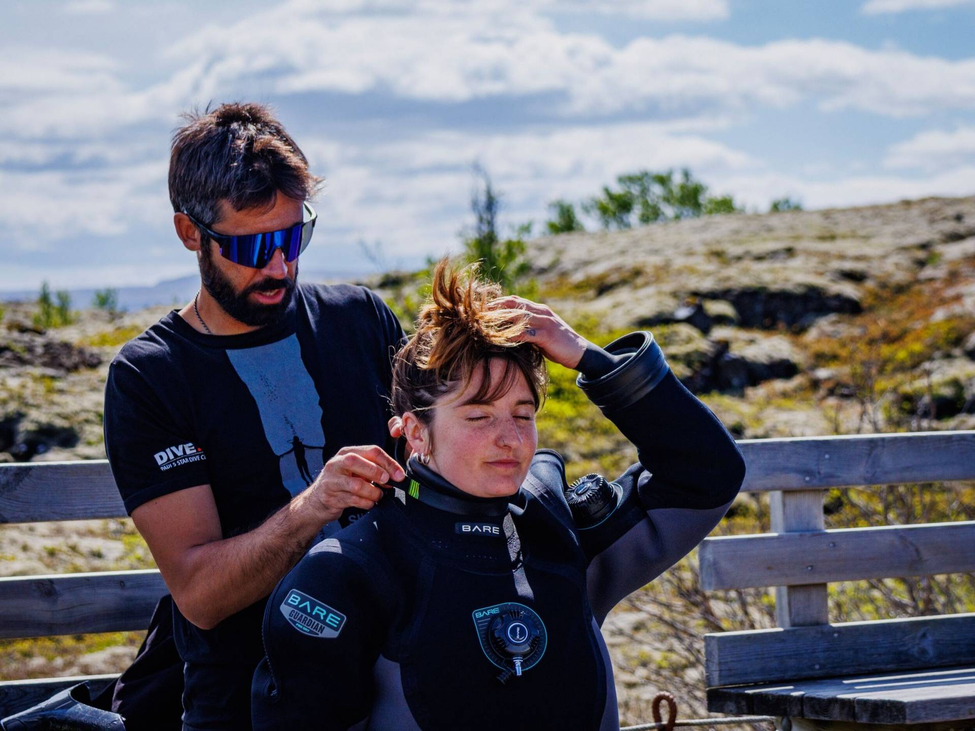 A male diver assisting a female diver with the straps on the dry suit